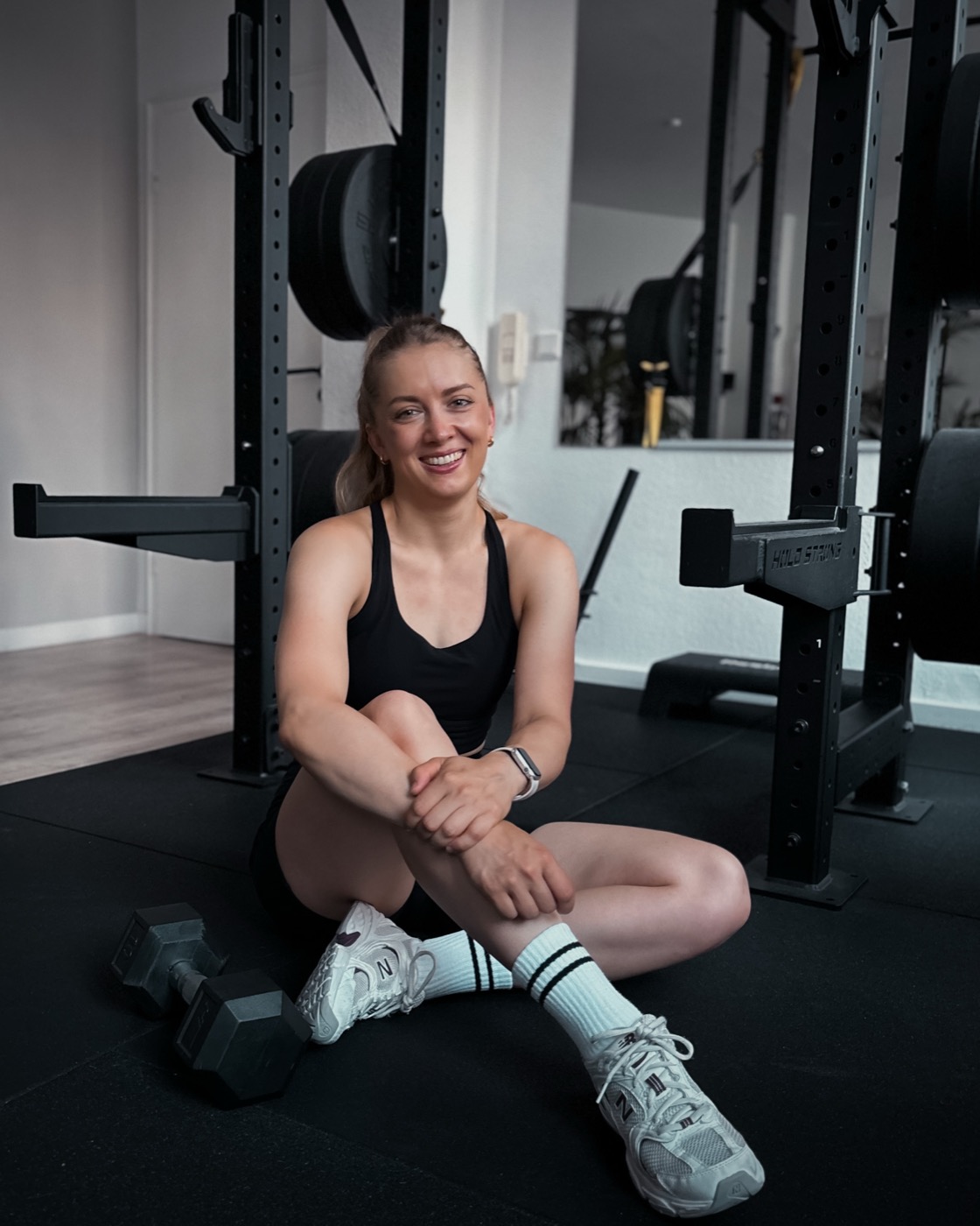 Julia Reinhardt sitzt lächelnd auf dem Boden ihres Personal-Training-Gyms vor einem Power Rack